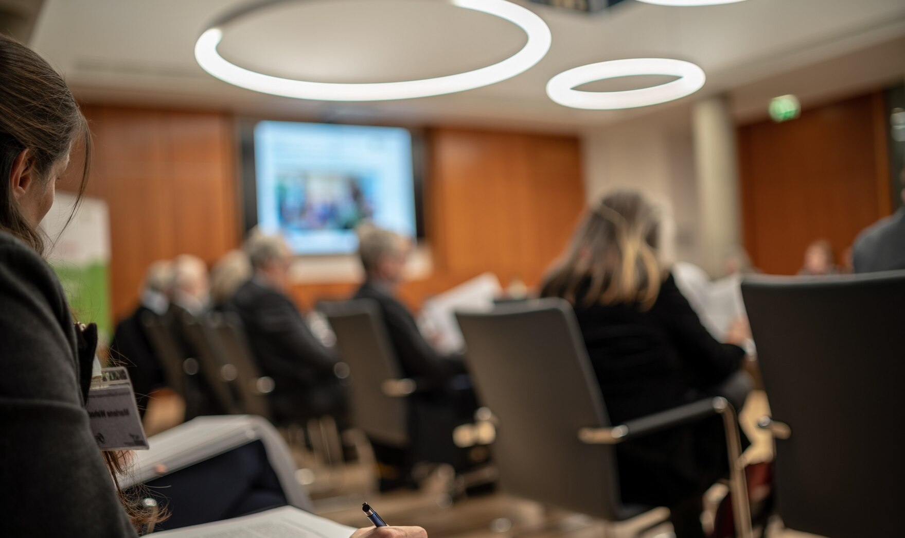 Die Mitglieder der Arbeitsgruppe sitzen an einem runden Tisch, im Hintergrund ist ein Leinwand auf grünem Untergrund zu sehen.