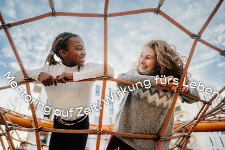 zwei Mädchen auf dem Spielplatz liegen in einem Kletternetz und schauen sich lächelnd an.