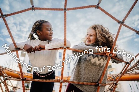 zwei Mädchen auf dem Spielplatz liegen in einem Kletternetz und schauen sich lächelnd an.