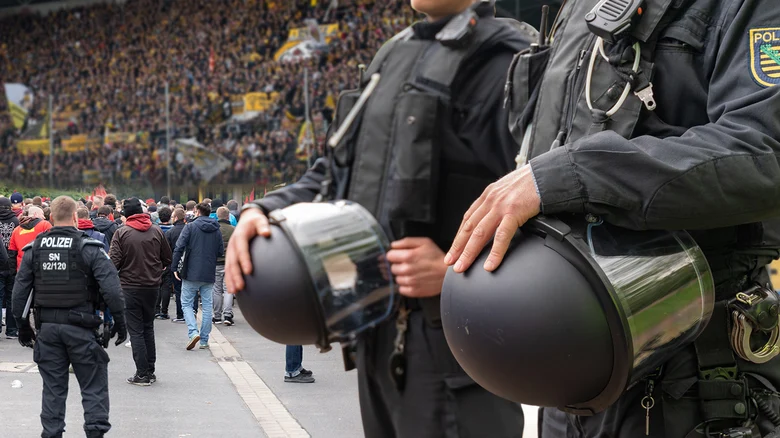 zwei Polizisten in dunkelblauer Einsatzuniform halten ihre Schutzhelme in der Hand. Sie stehen in einem Fußballstadion und im Hintergrund sitzen die Fans. Einige laufen auch noch, um ihre Plätze zu erreichen.
