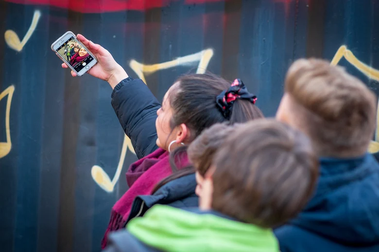 eine junge Frau hält in einer Hand ein Selfie und dahinter stehen drei weitere Jugendliche die alle in das Handy schauen und lächeln