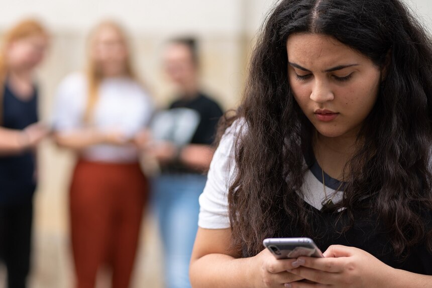 eine Jugendliche im Vordergrund schaut auf ihr Handy im Hintergrund sind drei weitere Jugendliche zu sehen, die ebenfalls auf ihr Handy schauen.