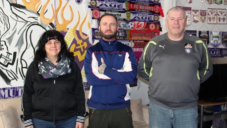 drei Personen, eine Frau links und zwei Männer neben ihr, lächeln vor einer Graffiti-Wand in die Kamera.