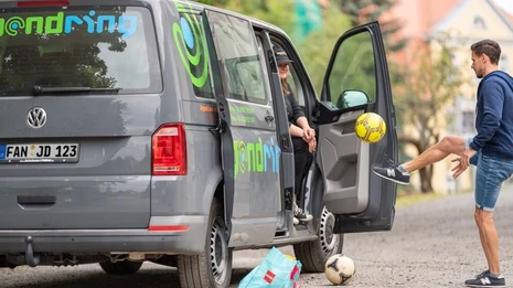 ein kleiner Tansporter mit geöffneter Schiebetür, auf dem Beifahrersitz sitze eine Person mit Basekap, die einer anderen Person, die vor der Tür steht einen gelben Ball zuspielt.