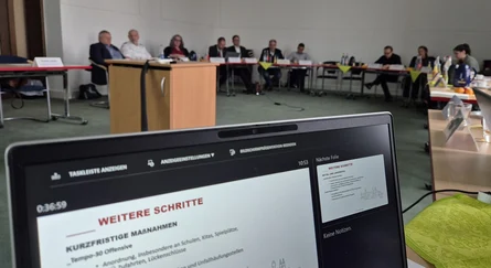 ein Ausschnitt von einem Laptop-Bildschirm dahinter verschwommen Personen die an einem Tisch auf Stühlen sitzen