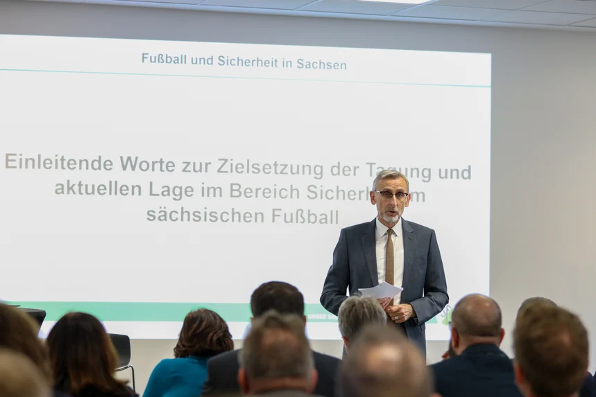 Innenminister Armin Schuster begrüßt die Teilnehmenden. Er steht vor einer Leinwand und spricht