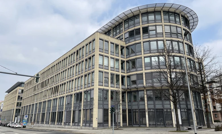 Außenansicht eines länglichen, mit Glasfenstern ausgestattetem beigem Gebäude. Davor eine Straße mit kargen Bäumen.