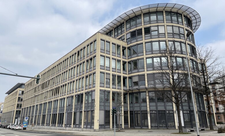 Außenansicht eines länglichen, mit Glasfenstern ausgestattetem beigem Gebäude. Davor eine Straße mit kargen Bäumen.
