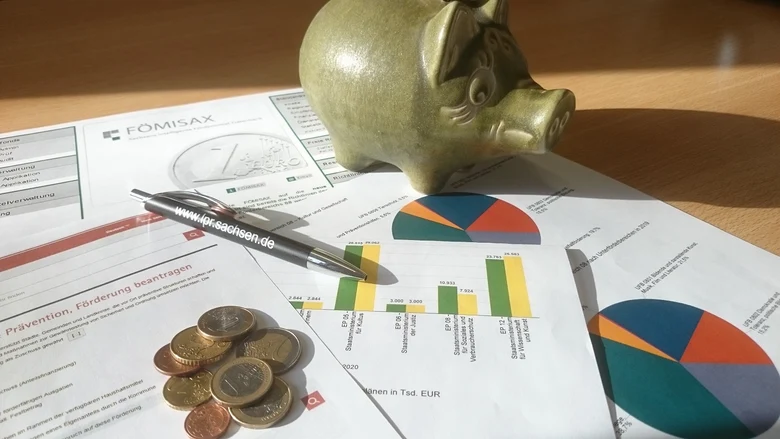 Auf einem Schreibtisch liegen mehrere Blätter mit bunten Kreis- sowie Balkendiagrammen. Ein goldenes Sparschwein, ein schwarzer Kugelschreiber und ein kleiner Haufen Münzen liegen darauf.