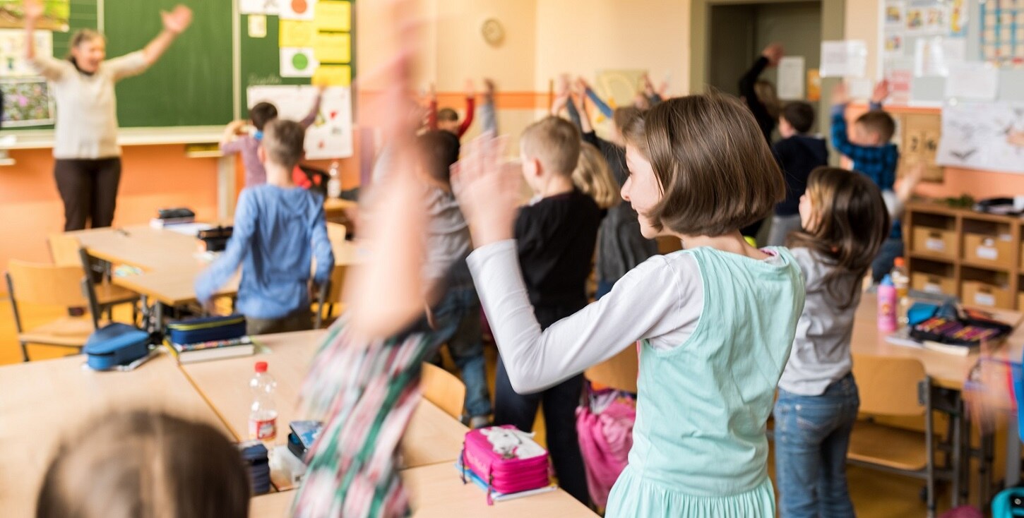 Viele Schulkinder in einem Klassenzimmer machen tanzende Bewegungen. An der Tafel vorn ist eine Lehrerin zu sehen.