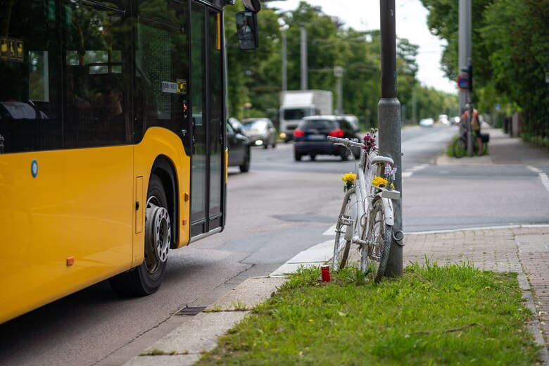ein weißes Fahrrad lehnt an einer Laterne neben einer Straße, auf der Straße steht ein gelber Bus des Personennahverkehrs. An dem Fahrrad sind Blumen befestigt.