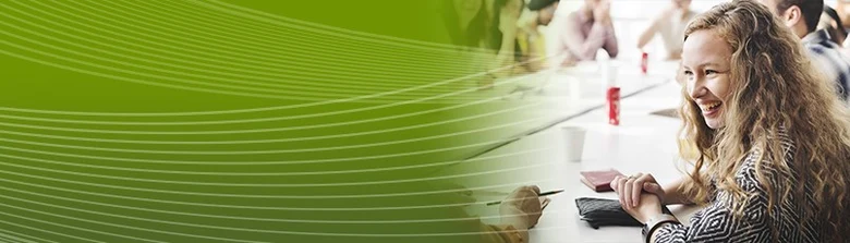 Werbebild für das Bachelorstudium »Präventionsmanagement«. Eine jungen Studentin, die sich fröhlich mit einer anderen Person unterhält. Im Hintergrund sitzen weitere Studierende an dem langen, weißen Tisch und tauschen sich aus.