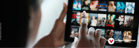 verschwommen ein Desktop mit verschiedenen Filmangeboten, davor sitzt eine Person, die mit dem Finger symbolisch einen Film anklickt