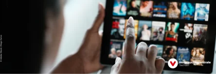 verschwommen ein Desktop mit verschiedenen Filmangeboten, davor sitzt eine Person, die mit dem Finger symbolisch einen Film anklickt