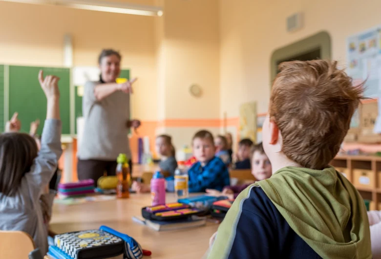 ein Klassenraum, Kinder sitzen auf Stühlen um einen Tische. Einige Schüler melden sich und der Lehrer, der vorn steht zeigt auf einen Schüler.igt auf einen Schüler