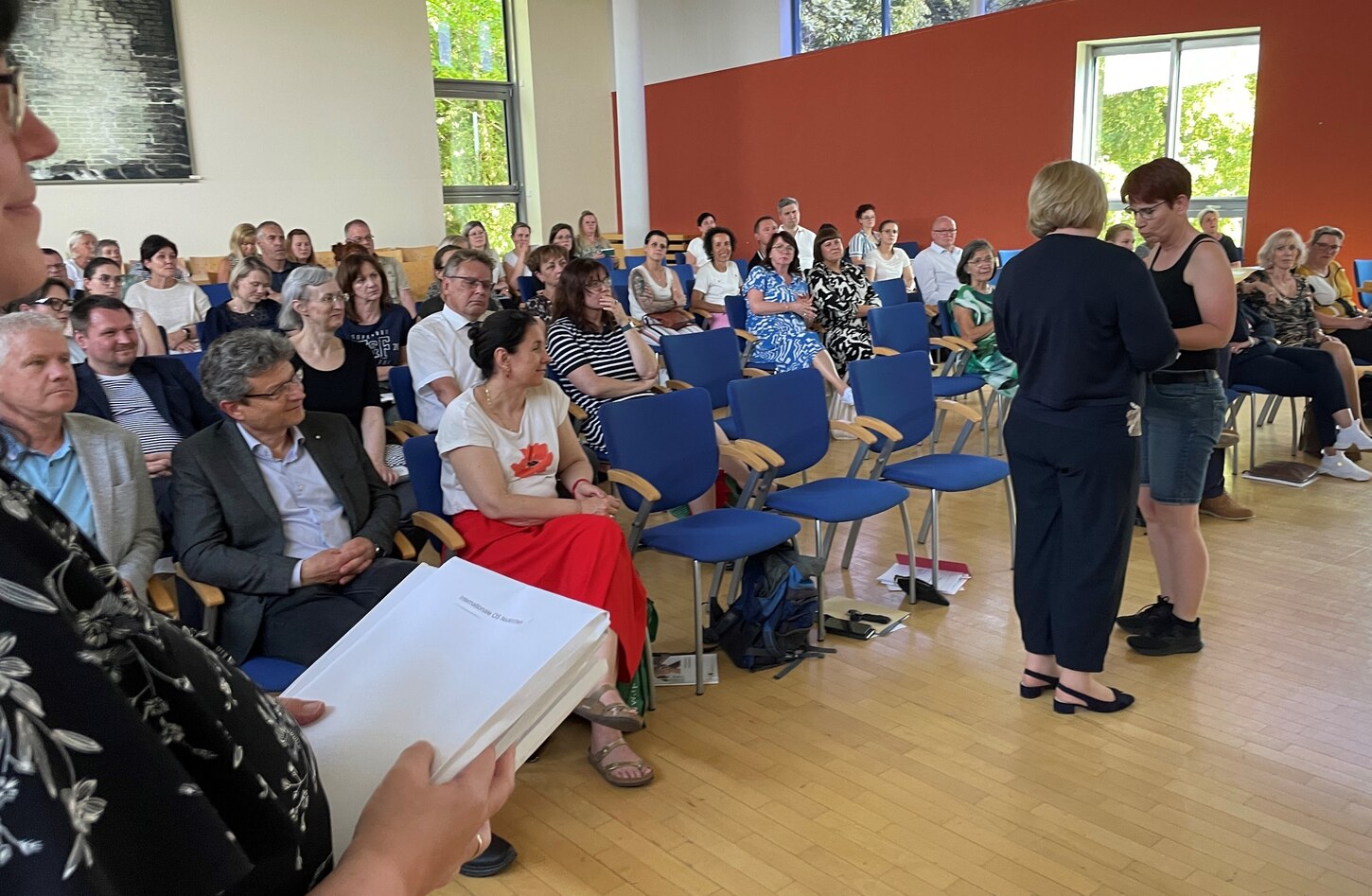Das Bild zeigt eine Versammlung von Menschen auf blauen Stoffstühlen sitzend. Auf der freien Fläche davor übergibt eine Frau ein Zertifikat.