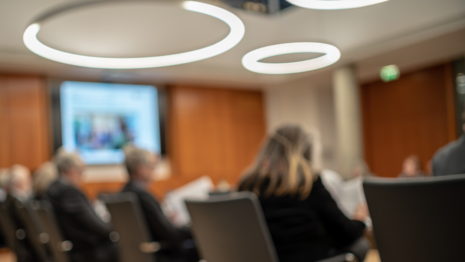 unscharfes Bild eines Konferenzraums, im Hintergrund mehrere Personen an einem Tisch sitzend 