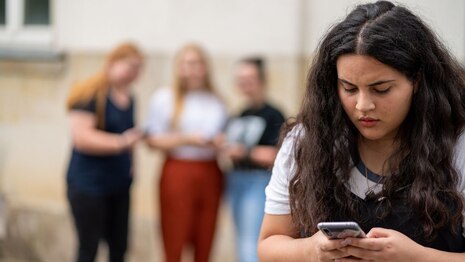 Gestellte Szene, ein Mädchen im Vordergrund schaut traurig auf ihr Smartphone, dahinter etwas verschwommen drei Mädchen, die sich über sie lustig machen.