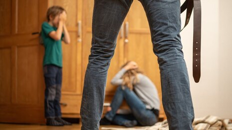 eine Frau kauert vor einem Schrank, links neben ihr ein Junge der sich die Augen zuhält und davor sind Beine eines Mannes zu sehen und eine Gürtel, das Bild ist nachgestellt und soll häusliche Gewalt nachstellen