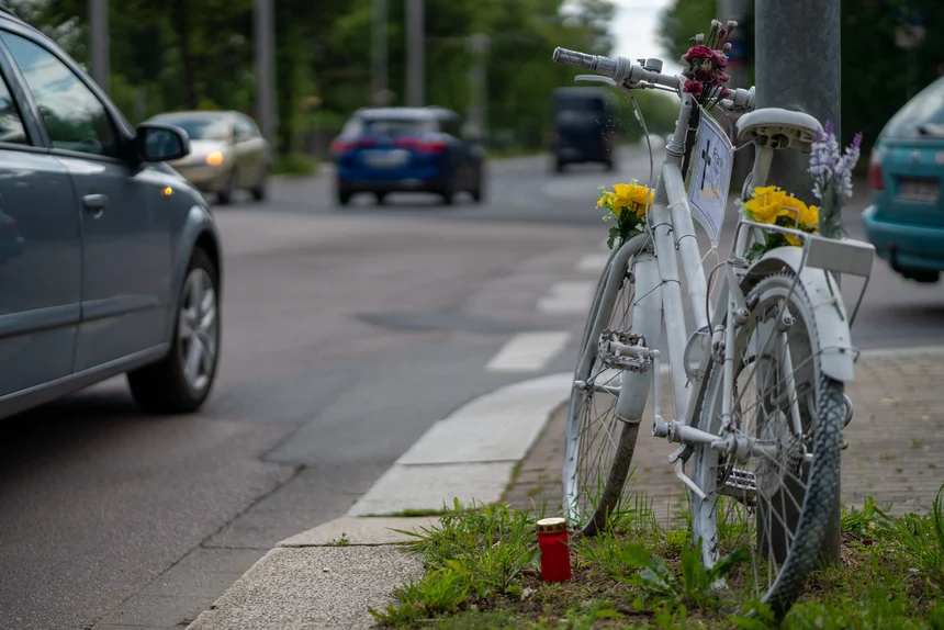 Ein weiß angemaltes Fahrrad geschmückt mit bunten Blumen an einer viel befahrenen Straße. Daneben steht eine rote Grabkerze für ein Unfallopfer.