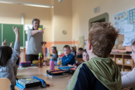 ein Lehrer steht vor der Klasse und zeigt auf einen Schüler, der wie viele andere Lernende in einem Klassenraum sitzt