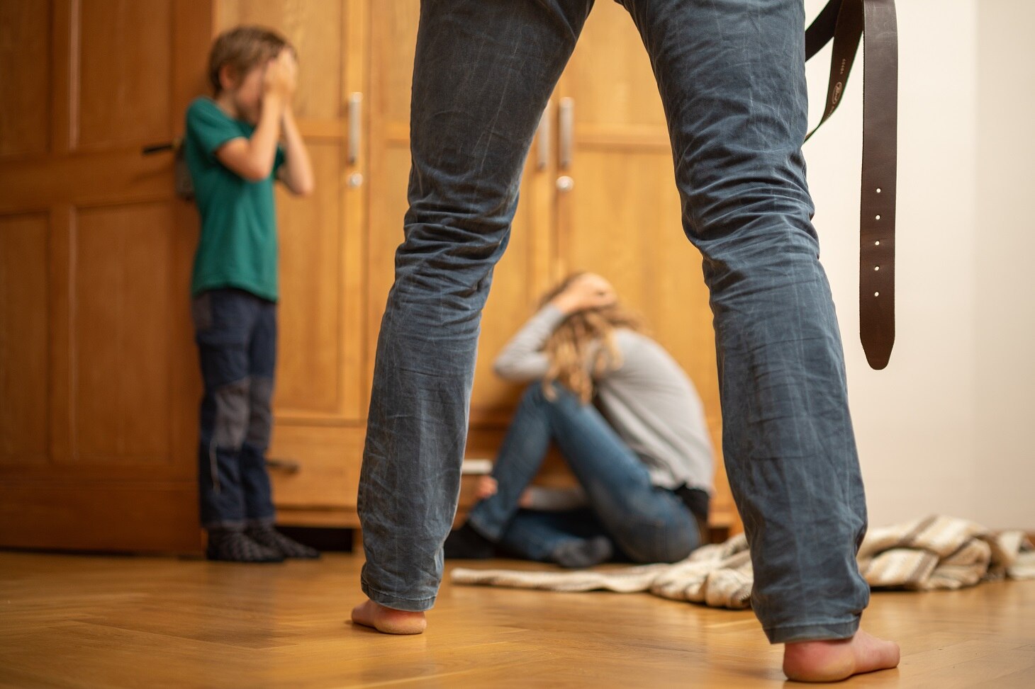 Gestellte Szene, häusliche Gewalt, ein Mann mit einem Gürtel in der Hand, eine Frau knieend auf dem Boden und daneben ein weinender Junge.